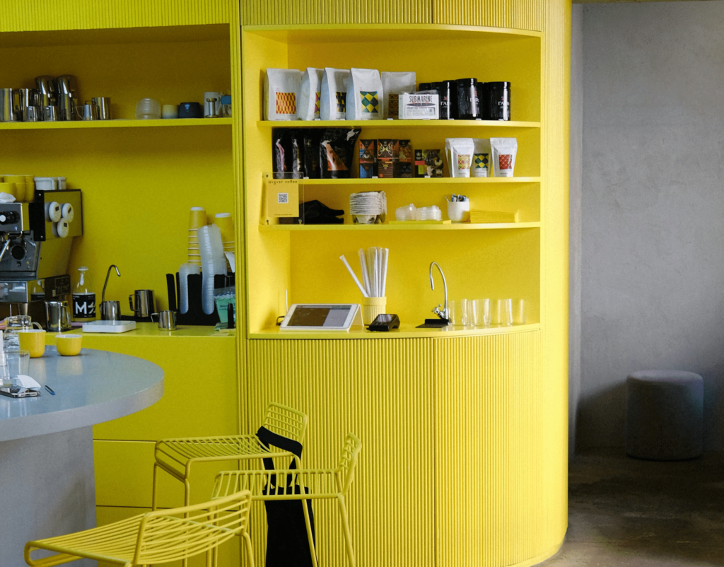 Coin café contemporain avec comptoir arrondi, chaises métalliques et étagères murales entièrement jaunes, mettant en valeur les produits et accessoires sur fond de béton brut.