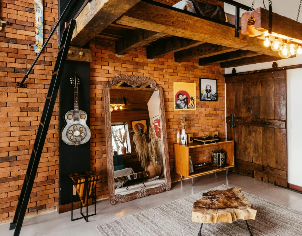 Salon de style industriel avec mur en briques, mezzanine en bois brut, table basse en tronc d’arbre, miroir sculpté, et échelle en métal noir, mélangeant matériaux naturels et design métal.