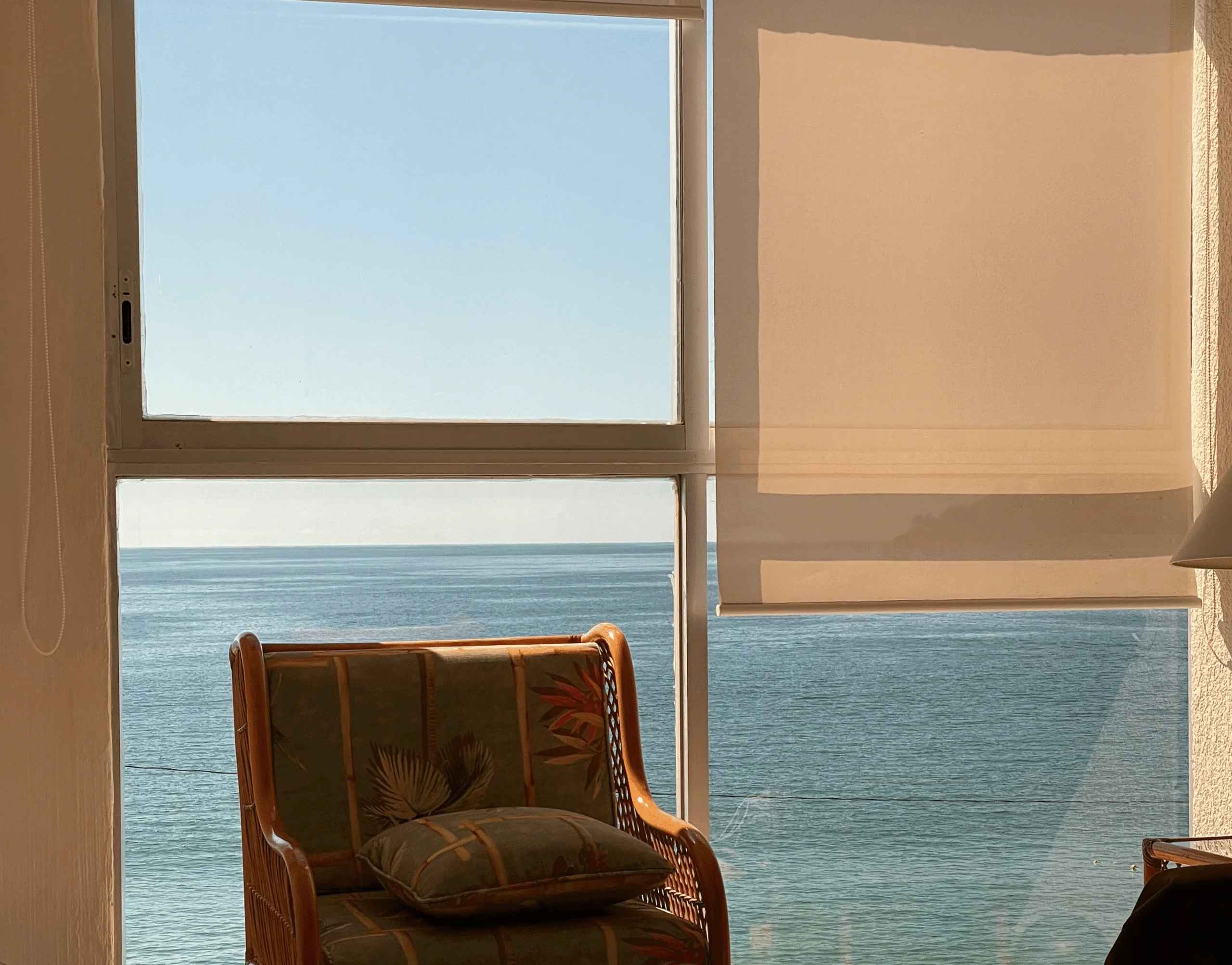 Fauteuil en rotin face à une grande fenêtre équipée de stores enrouleurs beiges, avec vue panoramique sur la mer et lumière naturelle filtrée.