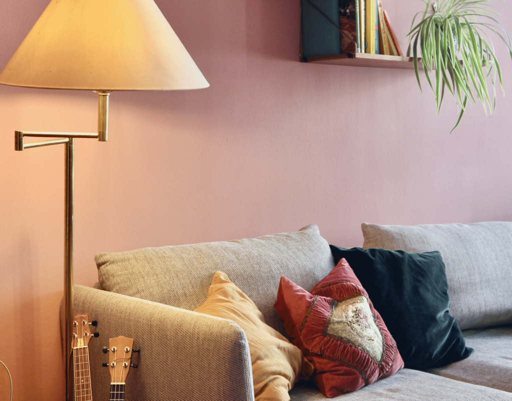 Salon avec canapé gris clair, coussins en velours et tissu brodé dans des teintes rose poudré, doré et vert foncé, mur rose mat en arrière-plan, lampe dorée et plante suspendue.